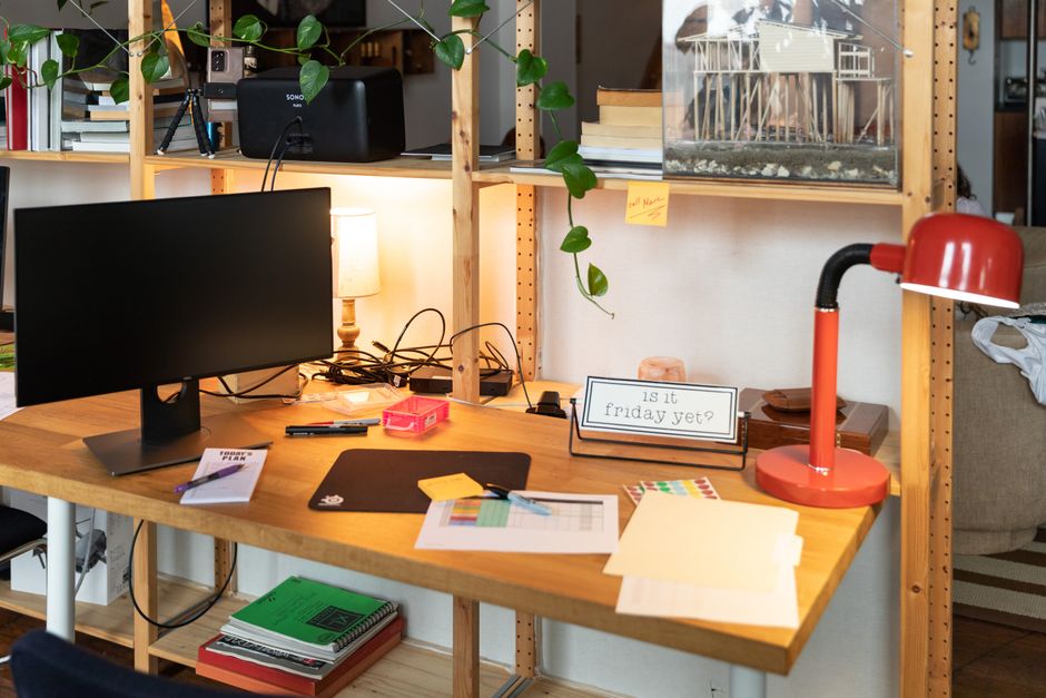 A cozy indoor workspace featuring a wooden desk, monitor, and red desk lamp, with plants and office supplies.